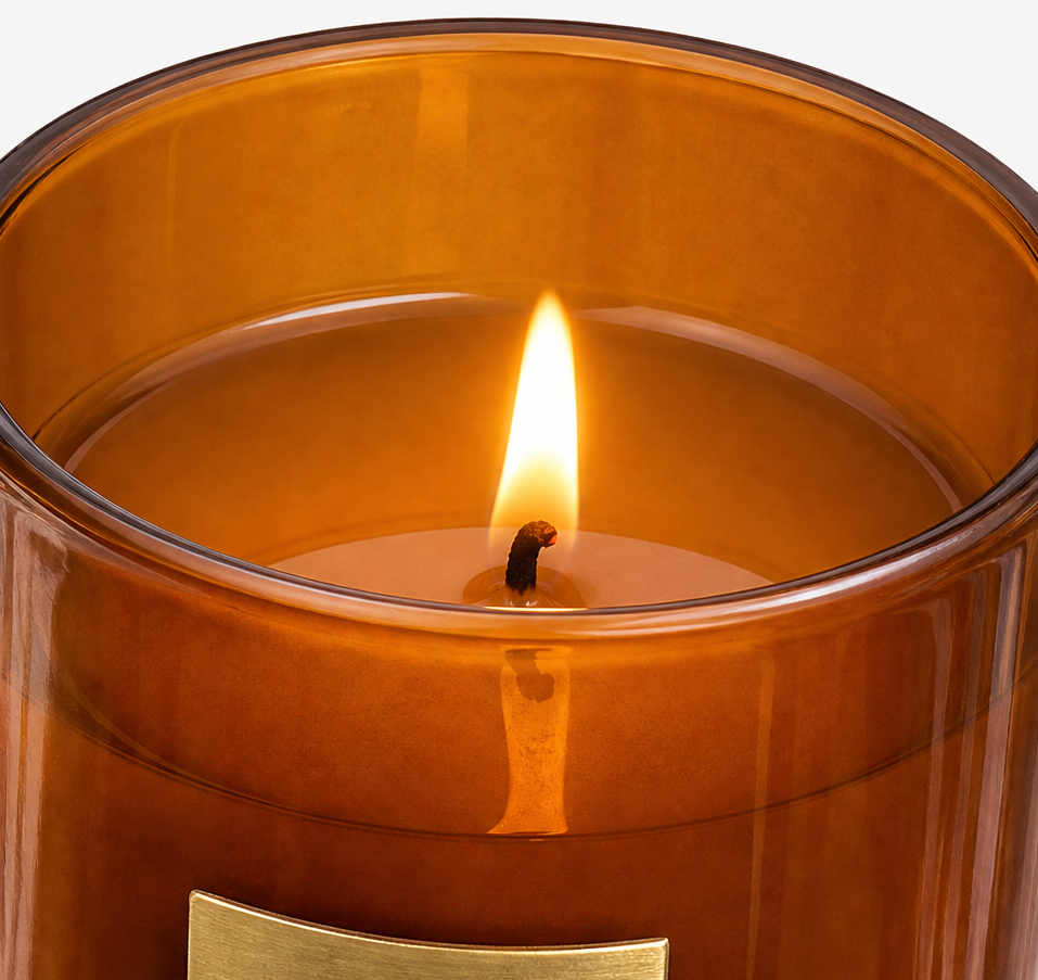 A close-up of a lit amber glass candle with a warm yellow flame and a small gold label at the bottom, set against a clean white background.