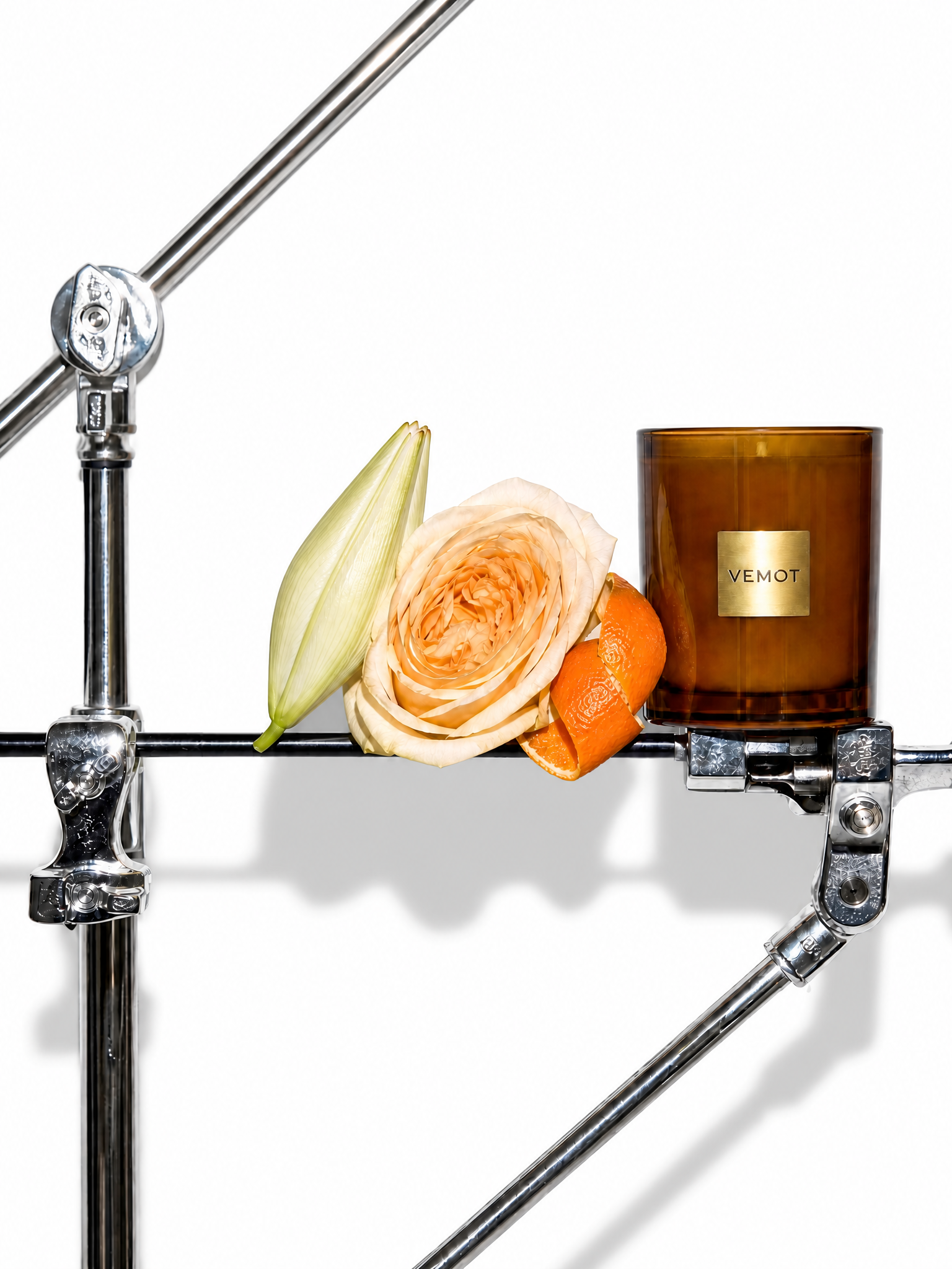 An amber glass candle jar with a gold VEMOT label, styled on a chrome metal stand with a peach rose, a pale lily bud, and orange peel against a white background.